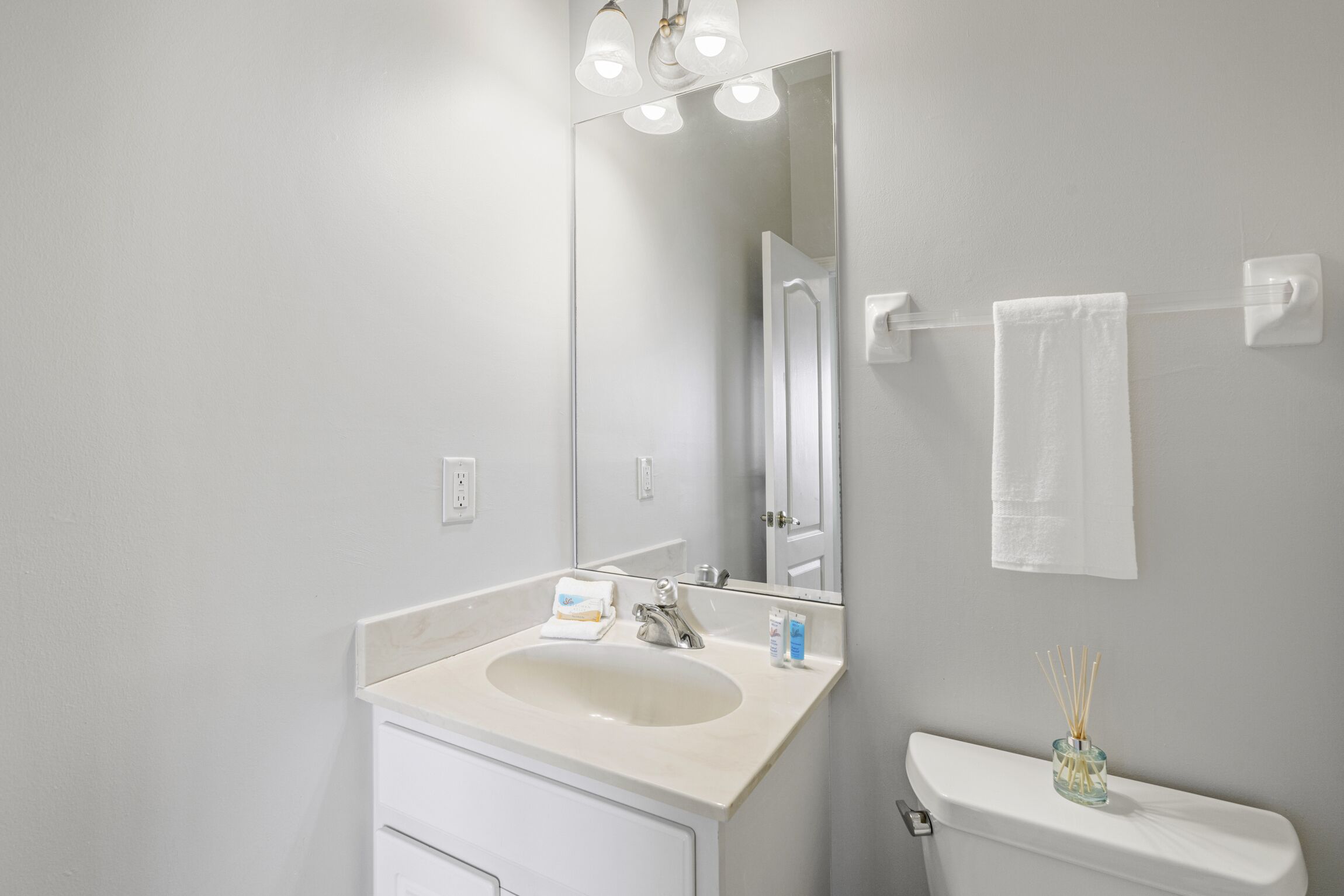 Une salle de bain avec un lavabo blanc, un miroir et des toilettes. Une serviette est accrochée au mur et un petit vase contenant des tiges de roseau est posé sur les toilettes.