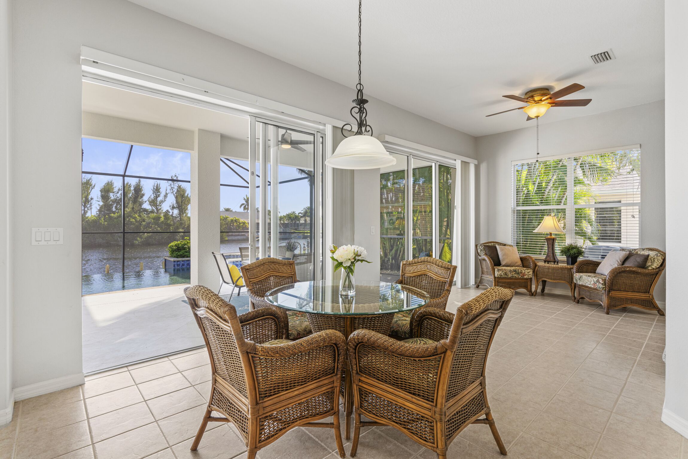 A spacious room with a glass table surrounded by wicker chairs, a ceiling fan, and a view of a water body through large glass doors.