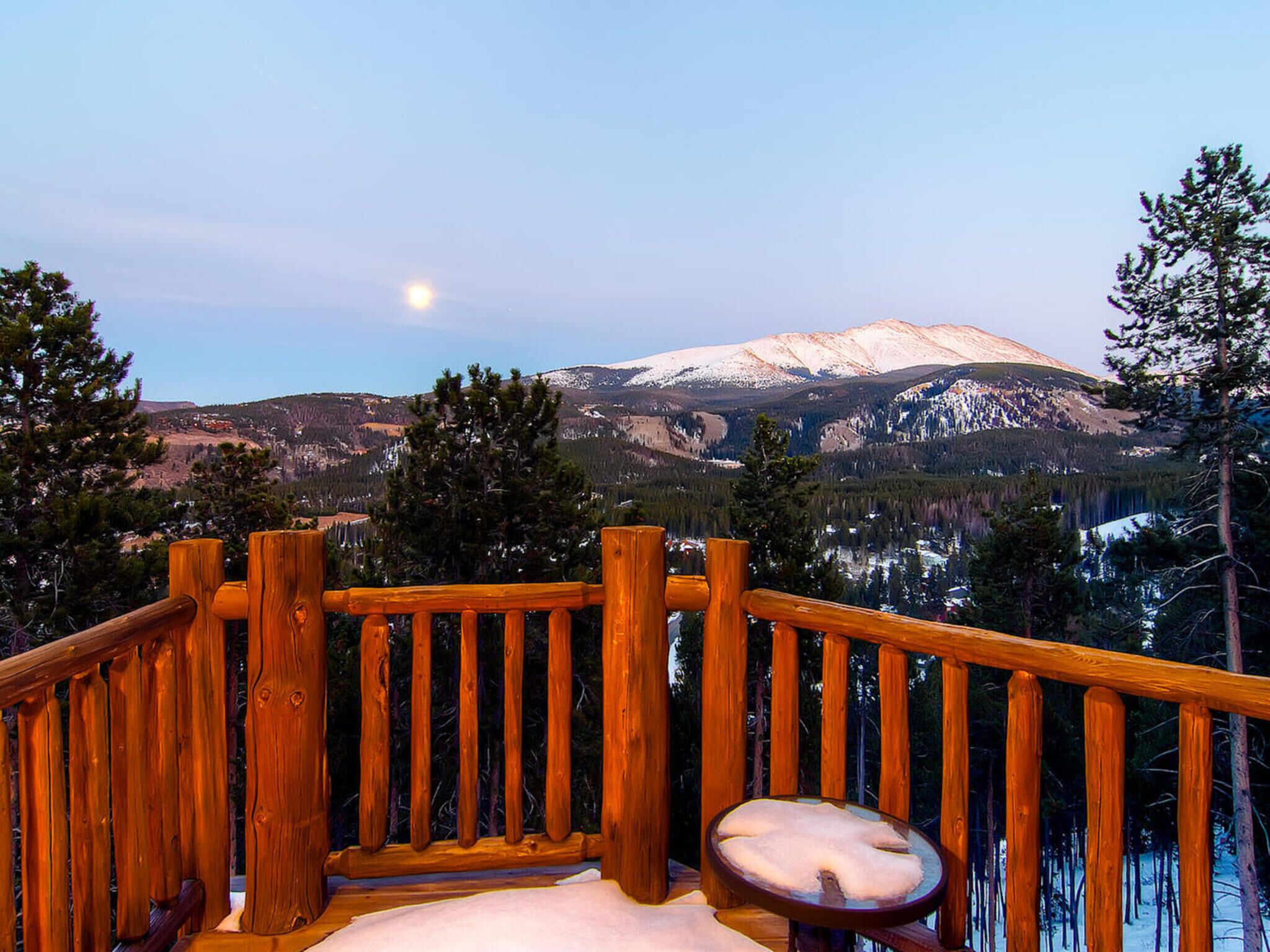 Una terraza de madera con un banco, con vista a una cadena montañosa cubierta de nieve.