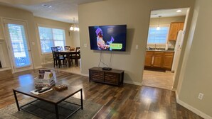 Uma sala de estar com televisão de tela plana, mesa de centro e área de jantar com cadeiras.