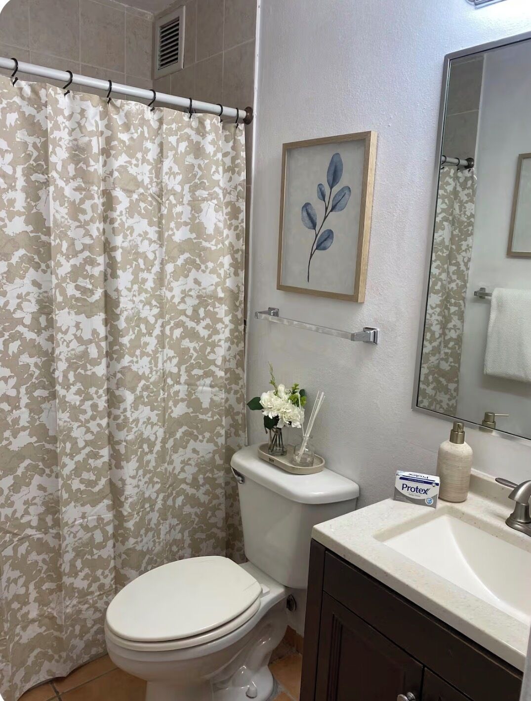 A bathroom with a camouflage shower curtain, a toilet, a sink with a Protex bottle, and a framed leaf artwork on the wall.