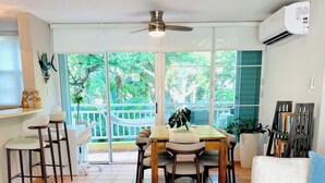 A living room with a sofa, a dining table with chairs, a ceiling fan, and a view of the outdoors through large windows. - Beachfront Condo+Pool+Backup Power+Full AC+FREEpkg+75TVSURR SYS (loiza)