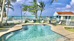 A swimming pool with a curved edge, surrounded by palm trees and a white fence. - Beachfront Condo+Pool+Backup Power+Full AC+FREEpkg+75TVSURR SYS (loiza)