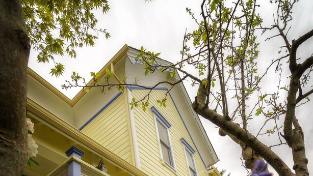 Une maison jaune à deux étages, dotée d’un balcon et d’un arbre au premier plan.
