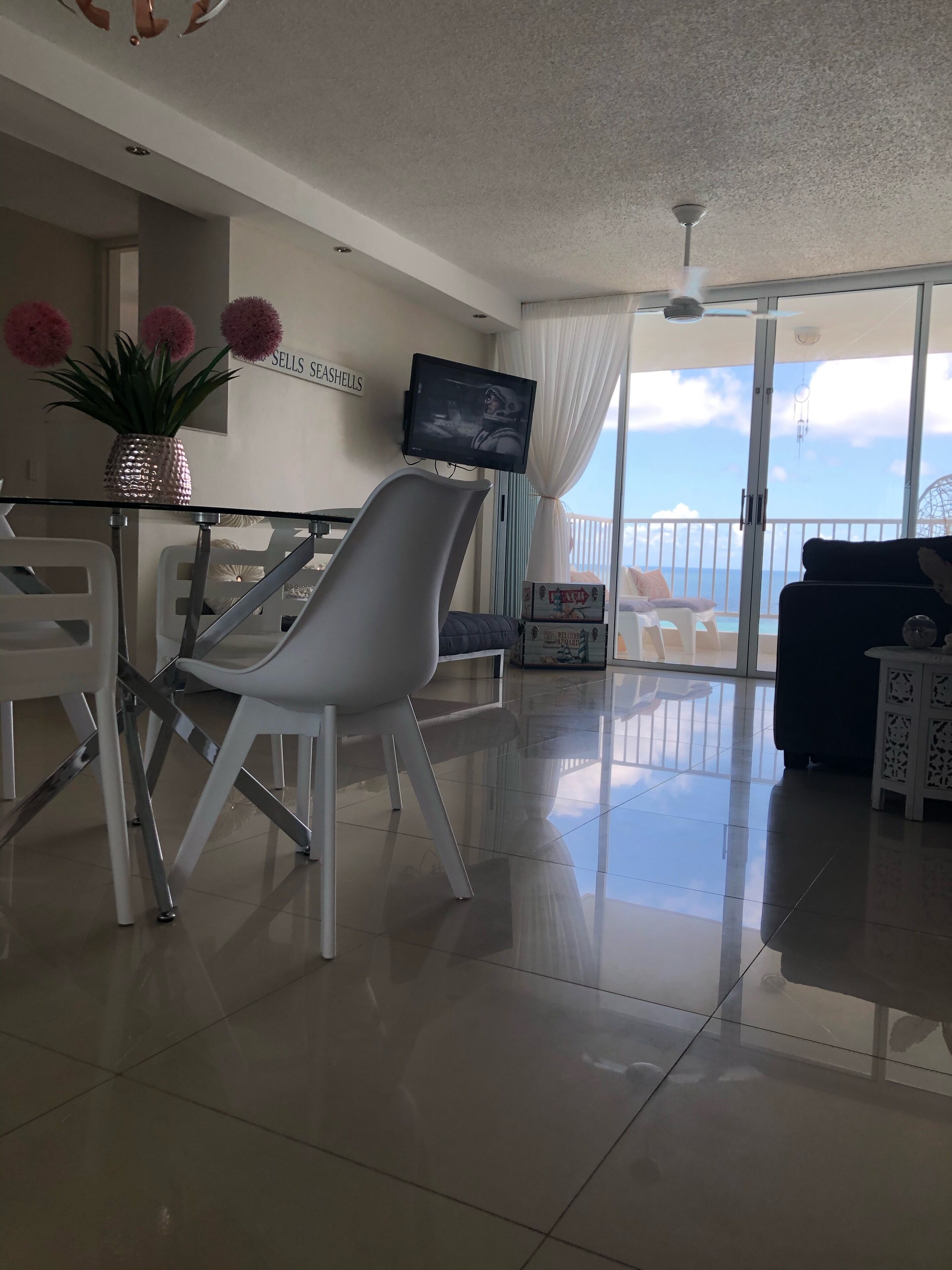 Ein modernes Wohnzimmer mit einem Glastisch, weißen Stühlen, einem Flachbildfernseher und Meerblick durch große Fenster.