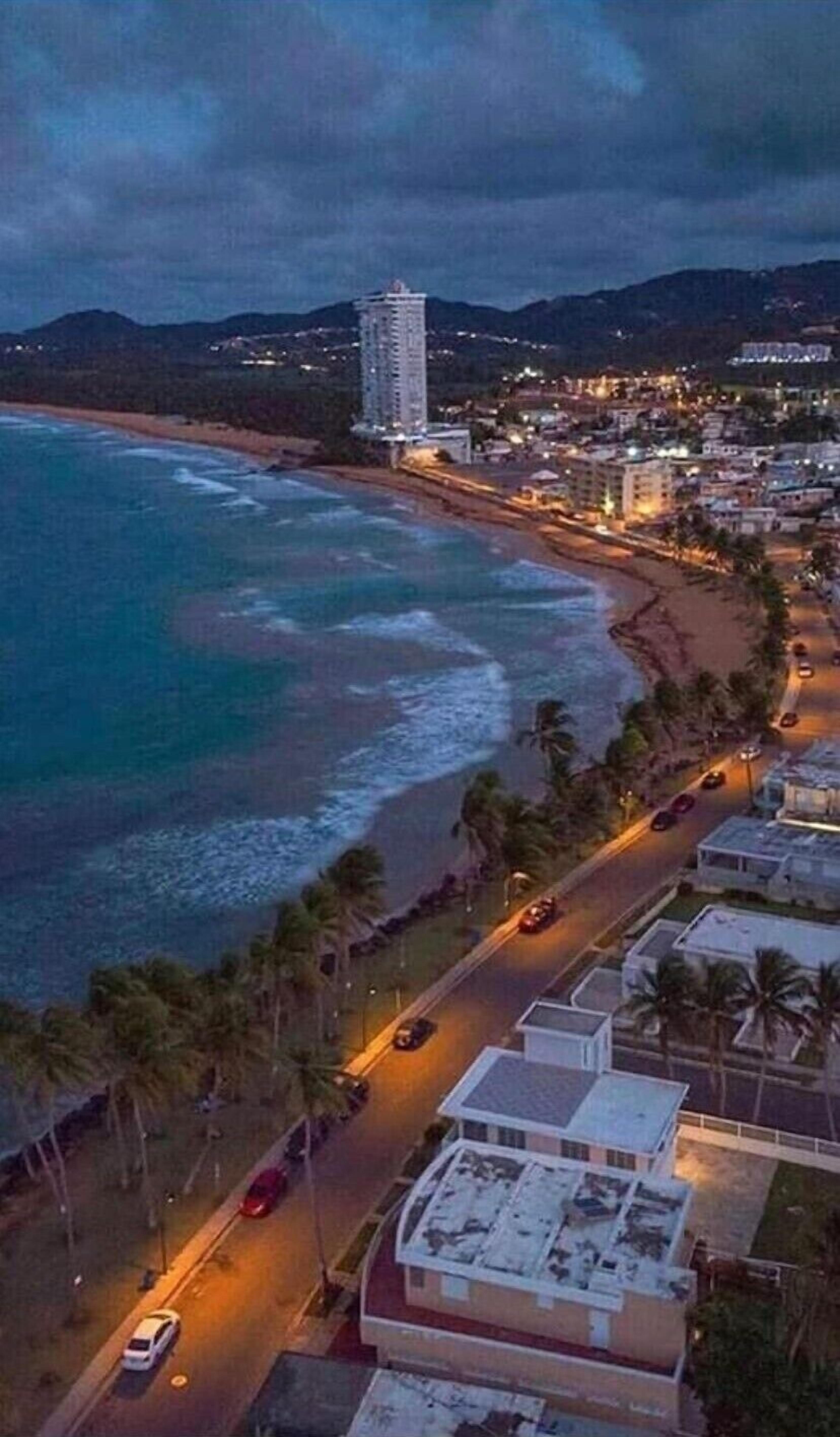 Un paysage urbain côtier avec une plage, un immeuble de grande hauteur et une route où circulent des voitures.