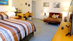 A hotel room with a bed, a sofa, a desk with a chair, a television, and a ceiling fan. - Wir sprechen Deutsch. Lovely studio with kitchenette, spacious lanai,TV and WiFi (Kailua Kona)