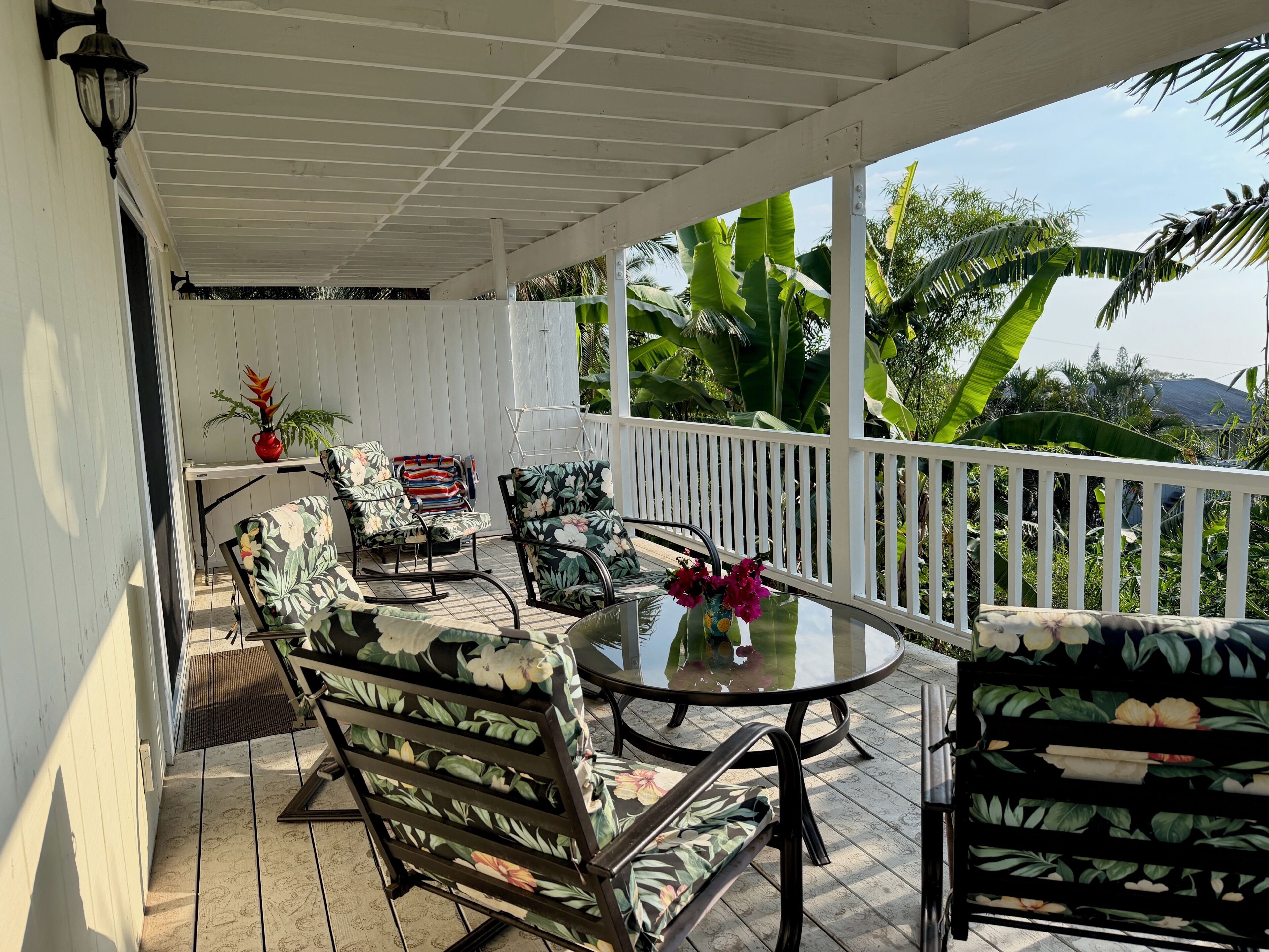 A covered outdoor seating area with a table and chairs, surrounded by lush greenery and a white railing.
