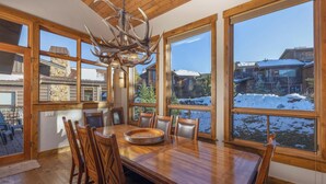 A dining area with a wooden table and chairs, large windows with a snowy landscape view, and a chandelier with antler decorations. - Retreat at Lewis Ranch: Walk to Lift, Private Hot Tub, Shuttle, Gas Grill (Copper Mountain)