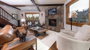 A cozy living room with a fireplace, a stone accent wall, and a large window overlooking a snowy landscape. - Retreat at Lewis Ranch: Walk to Lift, Private Hot Tub, Shuttle, Gas Grill (Copper Mountain)