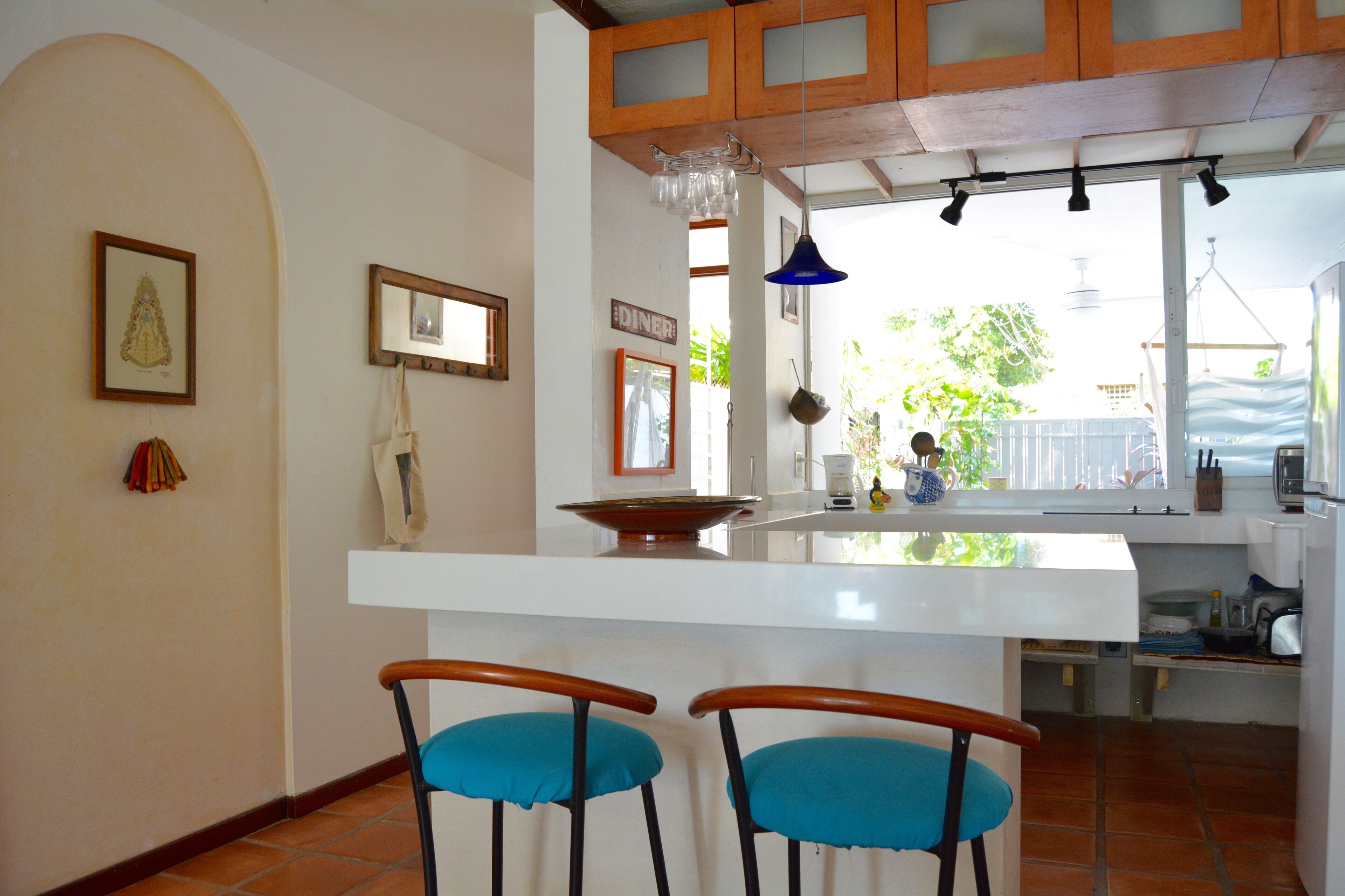 Une cuisine avec un îlot central blanc, deux tabourets de bar bleus avec assise rembourrée, des armoires en bois et un luminaire suspendu.