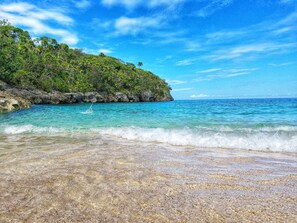 A beach with clear turquoise water, a rocky shoreline, and lush green vegetation on a hillside.