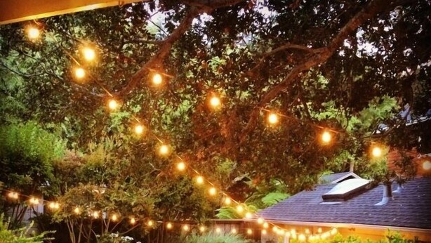 Un patio exterior con una mesa redonda y sillas, rodeado de abundante follaje y luces colgantes.