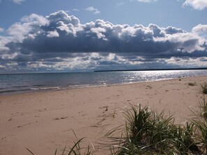 A beach with sand and grass in the foreground, a body of water in the middle ground, and a cloudy sky in the background.