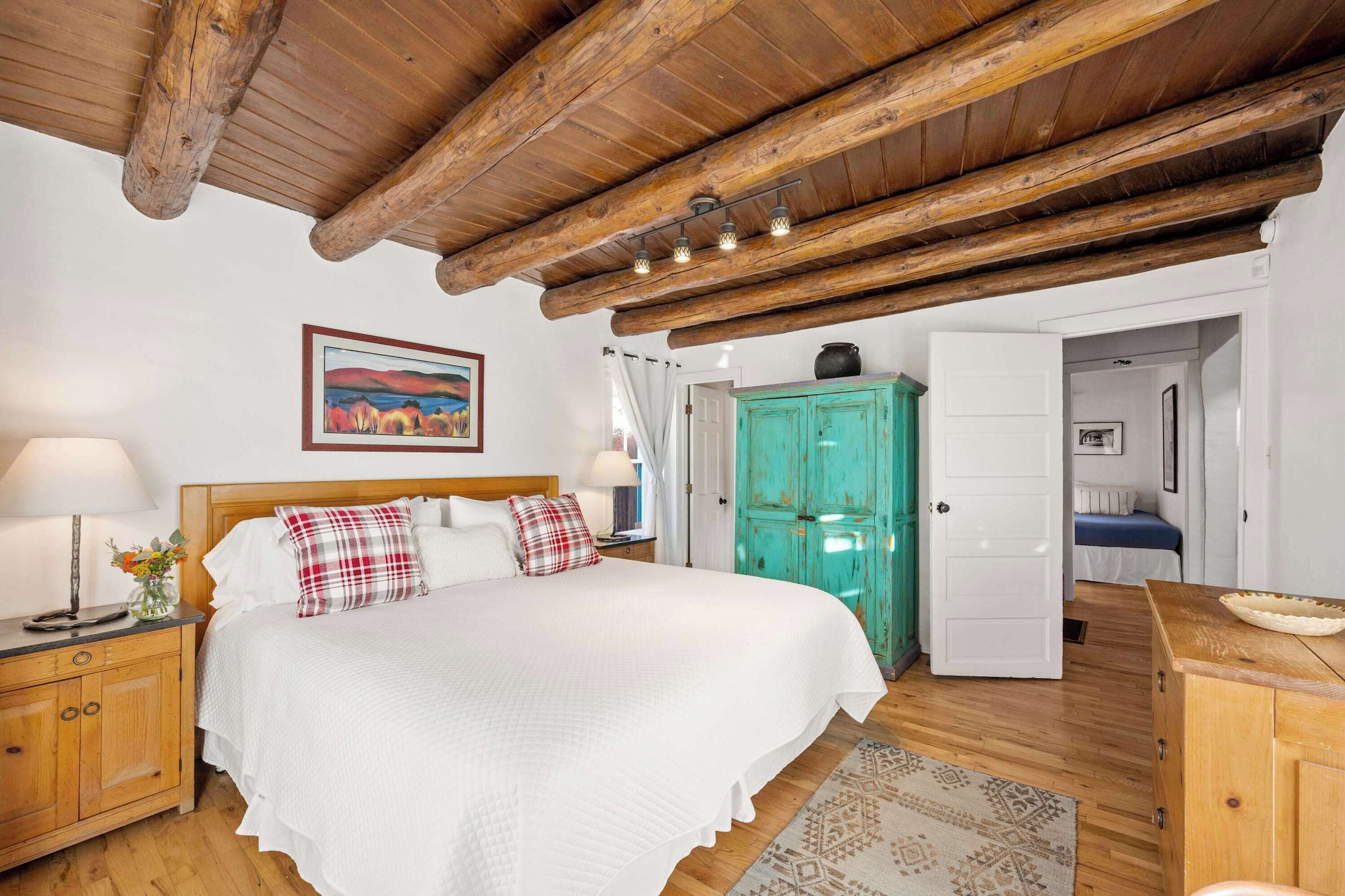 Une chambre avec un plafond en bois, un lit recouvert de linge de lit blanc, une commode turquoise et une table de chevet avec une lampe.