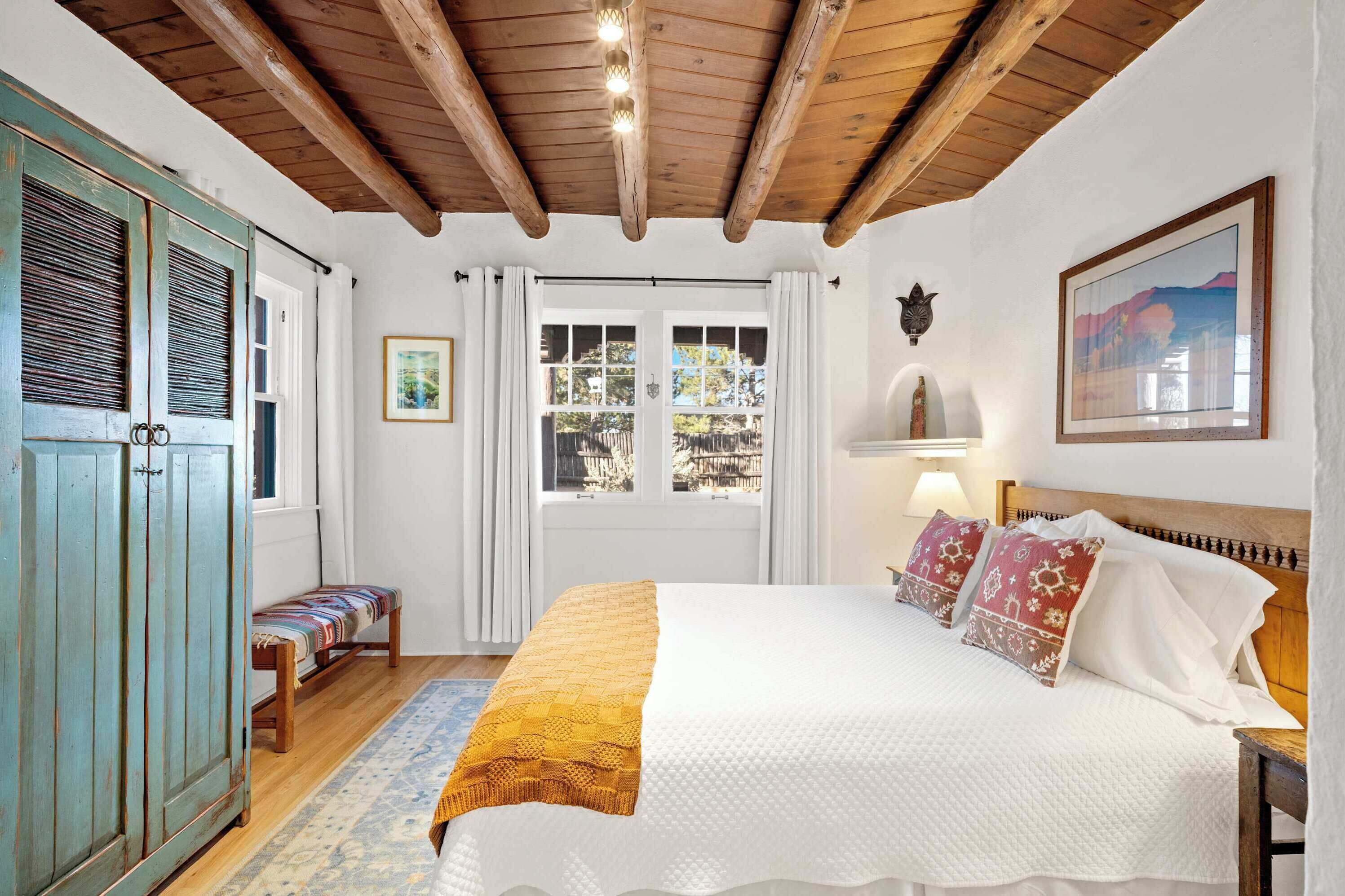 Un dormitorio con techo de madera, una cama con ropa de cama blanca y almohadas estampadas, un banco con un cojín de colores y una ventana con cortinas.