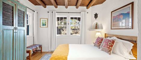 Un dormitorio con techo de madera, una cama con ropa de cama blanca y almohadas estampadas, un banco con un cojín de colores y una ventana con cortinas.
