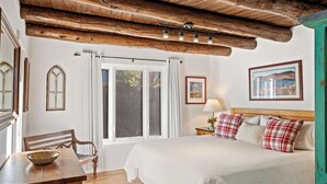 A bedroom with a wooden ceiling, a bed with white bedding, a nightstand with a lamp, and a window with curtains. - Historic Adobe, Walk to Railyard and the Plaza (Santa Fe)