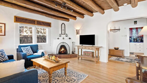 A living room with a fireplace, wooden beams, a television, and a kitchen area visible through an archway. - Historic Adobe, Walk to Railyard and the Plaza (Santa Fe)