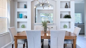 Un comedor con una mesa de madera y seis sillas grises tapizadas. Hay estanterías blancas con objetos decorativos y una lámpara de araña moderna sobre la mesa.