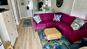 A compact living space with a purple sofa, a ceiling fan, a kitchen area, and a dining table. - Cutest Cottage on the Beach- FREE WIFI and Laundry Included sleeps 5 (Carolina Beach)
