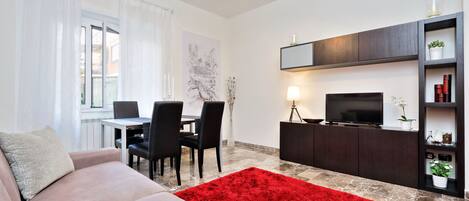 A modern living room with a sofa, dining table, chairs, a red rug, a TV on a cabinet, and a window with curtains.