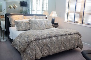 A bedroom with a large bed, patterned bedding, bedside lamps, and a window with blinds. - Oceanfront Bold & Stately Condo_Top Listing 2021 Books Early-Family Friendly (Myrtle Beach)