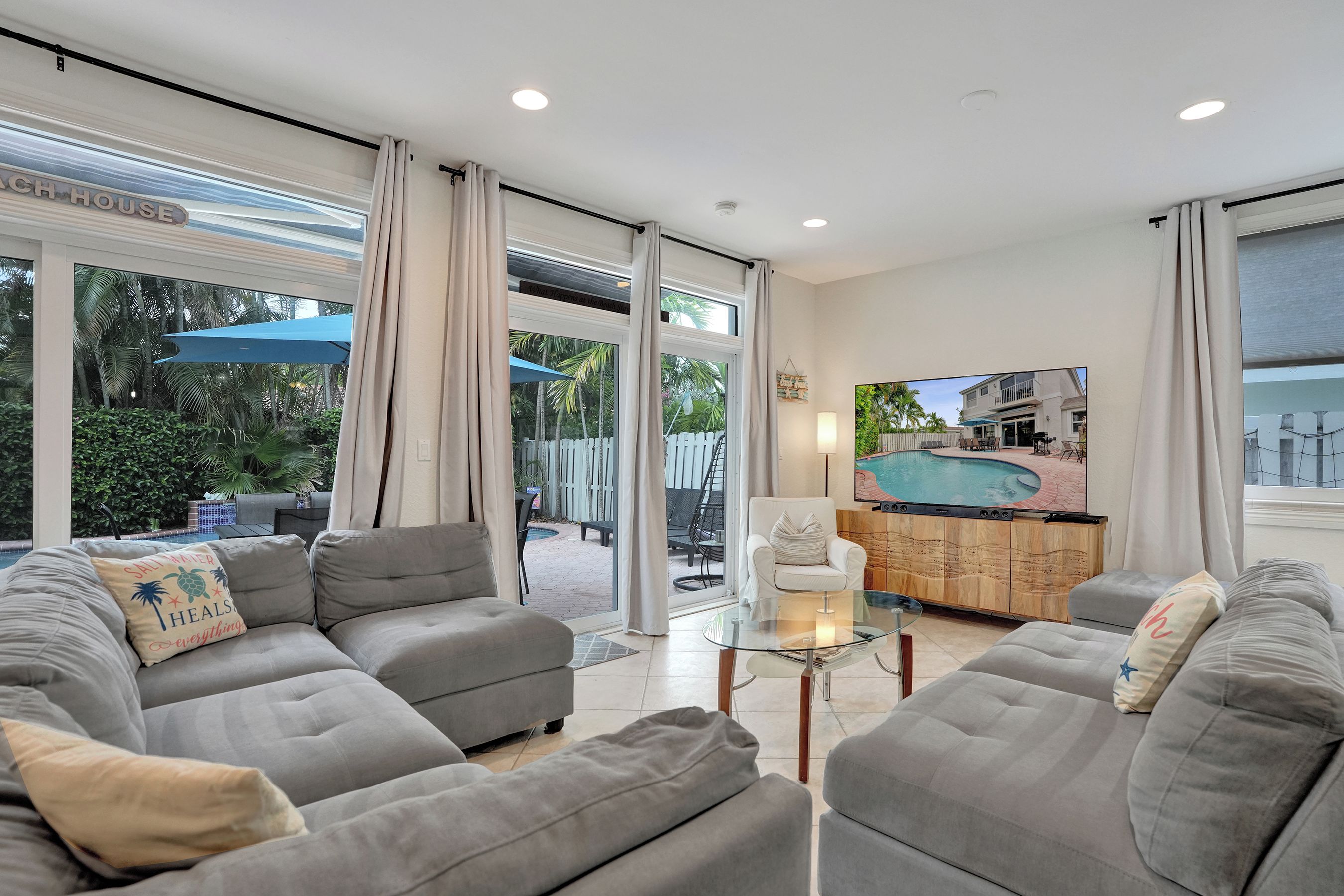 A modern living room with a grey sofa set, a glass coffee table, and a view of a pool and outdoor seating area.