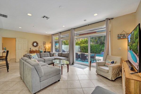 A spacious living room with a large gray sectional sofa, a glass coffee table, and a beige armchair. There is a television on a wooden stand to the right.