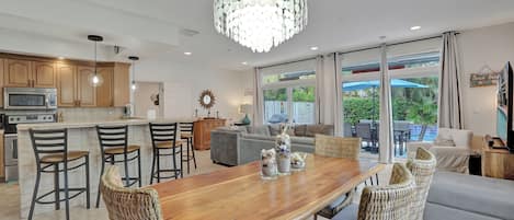 Cocina moderna con una isla central, comedor con mesa de madera y sillas de mimbre, y sala de estar con sofá y televisor.