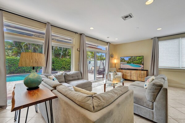 A spacious living room with a large sofa, a coffee table, and a TV. There is a view of a pool outside.