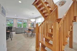 Un intérieur moderne avec un escalier en bois, un coin repas comprenant des chaises et une table, et de grandes fenêtres offrant une vue sur la verdure.
