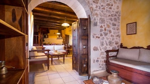 A rustic interior with a stone wall, wooden furniture, and an open archway leading to a kitchen area. - Purple villa Malotira - 1 bedroom -veranda -roof terrace - mountain and sea view (Chania)
