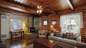 A log cabin interior with a stone fireplace, wooden furniture, and a ceiling fan.