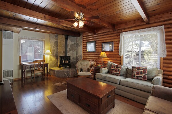 A log cabin interior with a stone fireplace, wooden furniture, and a ceiling fan.