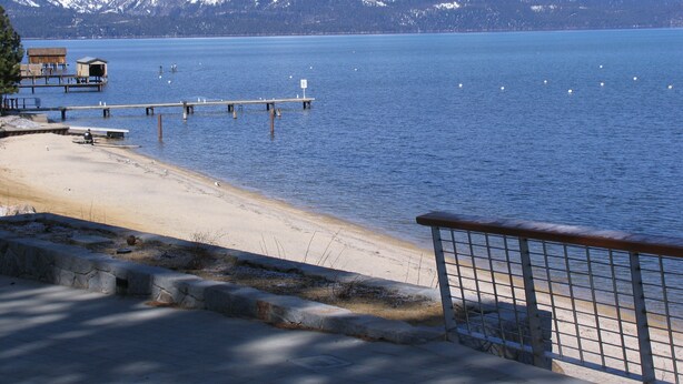 A lakeside view with a wooden pier extending into the water, a sandy beach, and snow-capped mountains in the background.
