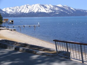 A lakeside view with a wooden pier extending into the water, a sandy beach, and snow-capped mountains in the background.