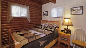 A wooden cabin room with a bed, a nightstand, a chair, and two windows with lace curtains.
