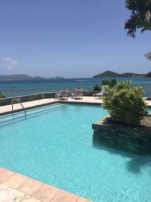 A pool with clear blue water, surrounded by lounge chairs and a stone wall.