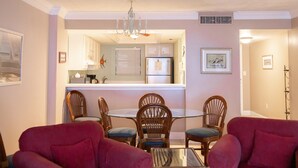 A dining area with a round table and six wooden chairs, a chandelier, and a kitchen in the background. - Oceanfront_Spacious_View the Sunrise_ Family Friendly & Quiet Building (Myrtle Beach)