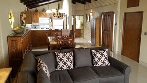 A living room with a dark grey sofa, patterned cushions, wooden dining table, and chairs. A kitchen is visible in the background with wooden cabinets and a white countertop.