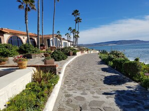 A coastal pathway lined with palm trees and potted plants, leading to a building with a tiled roof and arched windows. - (17#1 ) romantic and cozy condo in Las Gaviotas en Playas de Rosarito (Playas de Rosarito)