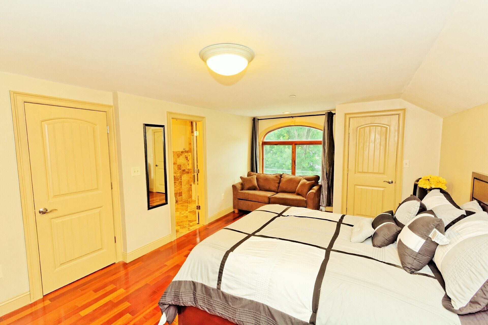 A bedroom with a large bed, a wooden floor, a window with a view of trees, and two doors.