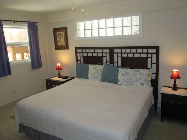 Una cama bien hecha con una colcha blanca, dos mesitas de noche con lámparas, una ventana con cortinas y un cuadro en la pared.