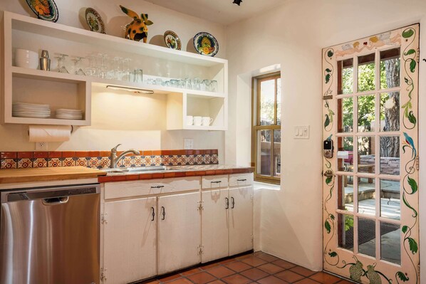 A kitchen with white cabinets, a stainless steel dishwasher, and a tiled backsplash. There are shelves above the sink holding plates and glasses. - Incredible Location, Hot Tub, Roof Deck (Santa Fe)