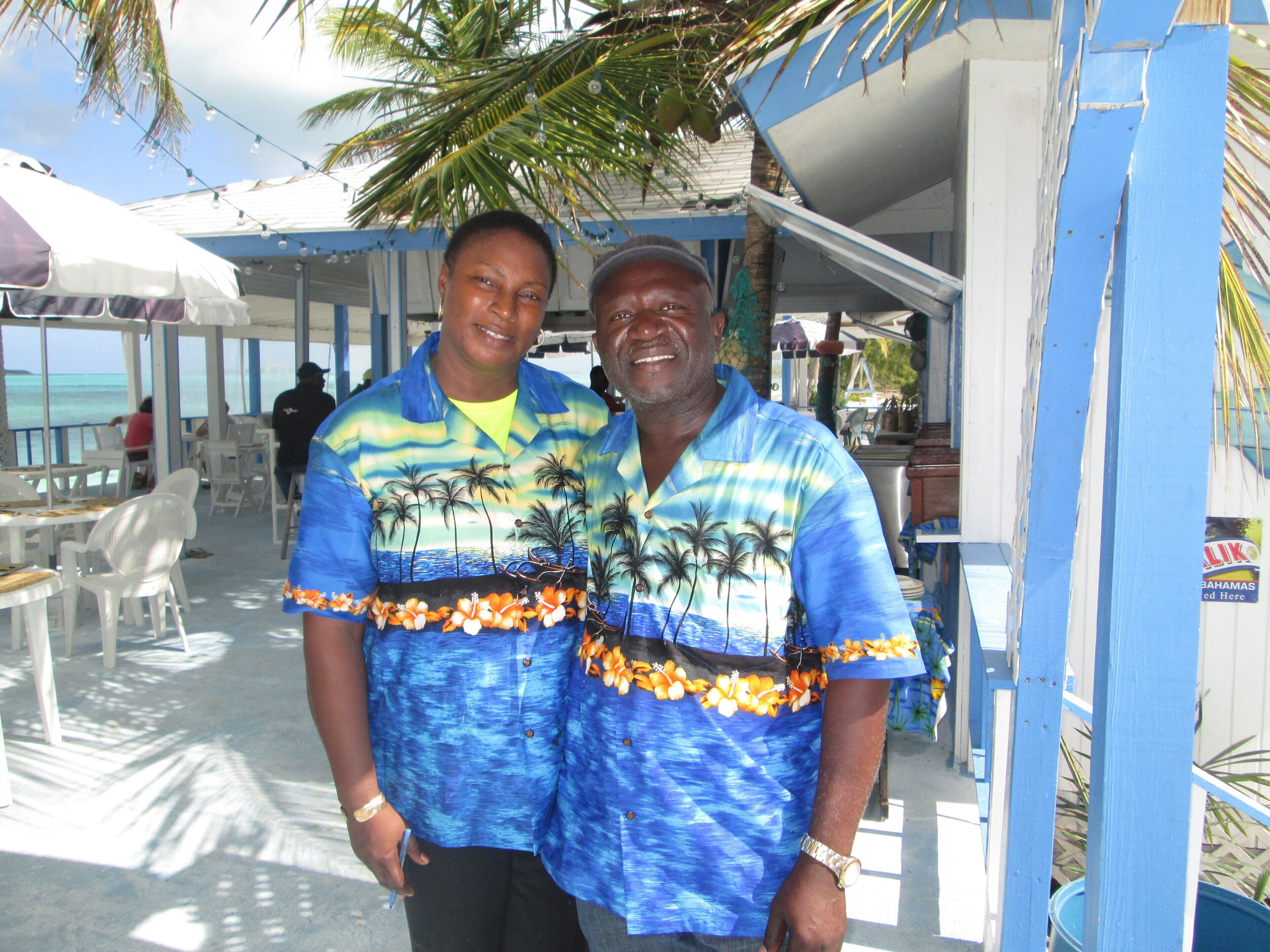 Two people standing in a covered outdoor area with tables and chairs, palm trees, and a view of the ocean.