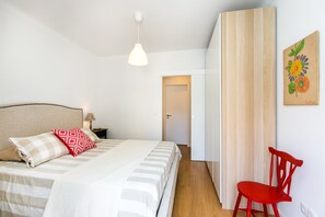 A bedroom with a bed, a nightstand, a red chair, a wooden door, and a wall painting.