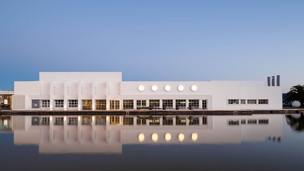 En moderne bygning med flad facade, store vinduer og et spejlbillede i vandet foran.