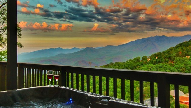 Un jacuzzi con dos copas de vino tinto, frente a un paisaje montañoso con un cielo colorido.