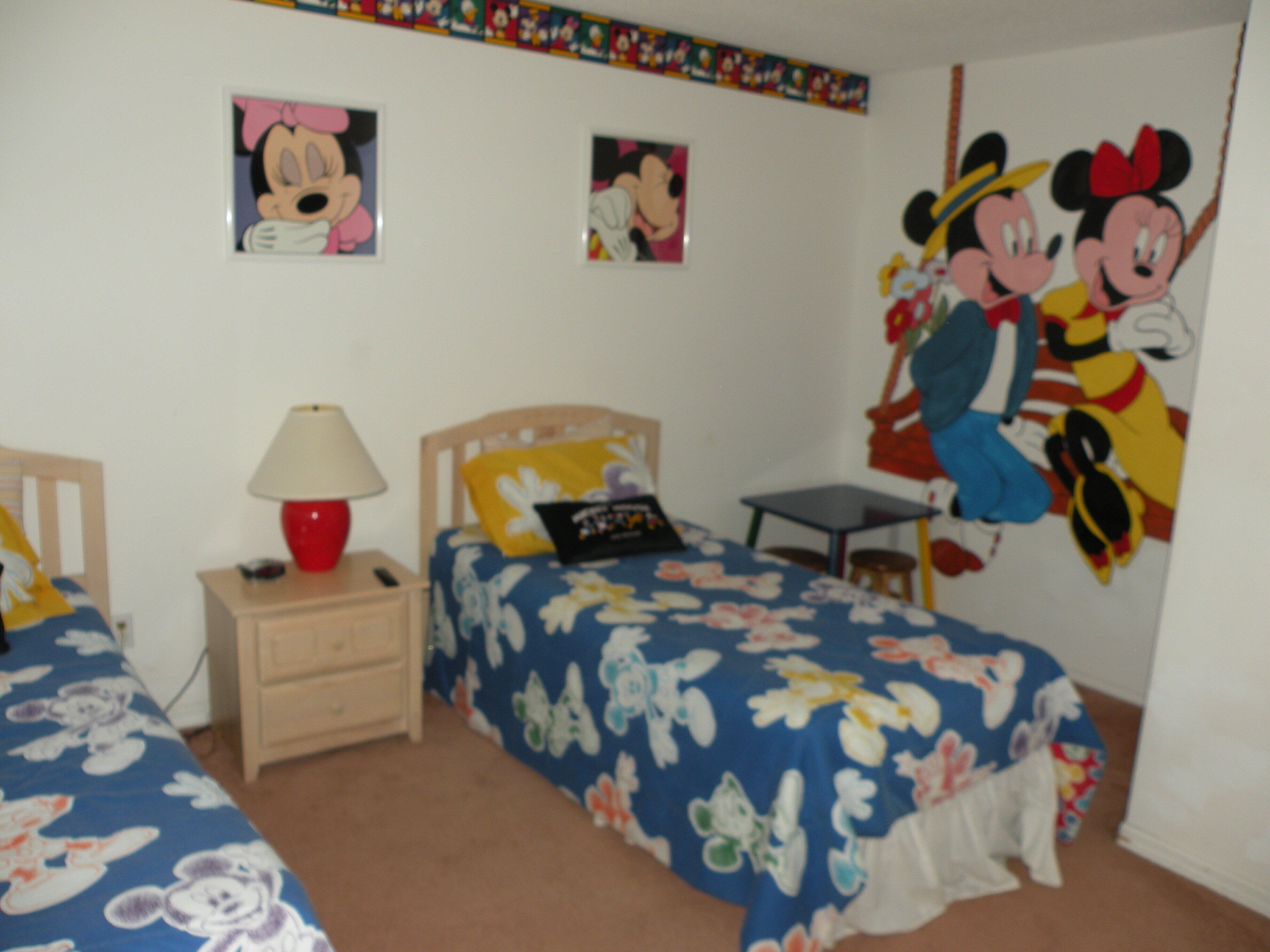 Ein Schlafzimmer mit zwei Betten, einem Nachttisch, einer Lampe und einem Mickey Mouse Poster an der Wand.
